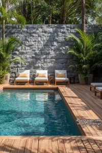 rectangular pool with wooden deck, plush loungers, stone privacy wall, and tropical plants. 5