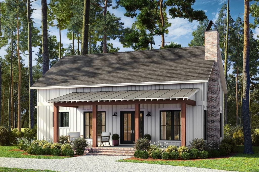 Front view of a 2-bedroom barndominium cottage house plan featuring a white board and batten exterior, brick chimney, covered porch with wooden posts, and surrounding landscaping, representing a 1,320 sq. ft. floor plan.