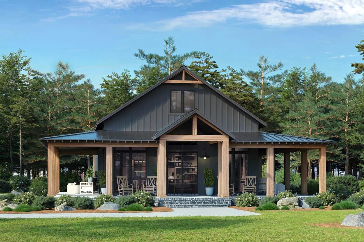 Front view of a 3-bedroom barndominium house plan featuring a wrap-around porch, dark metal roof, wooden columns, large windows, and surrounded by trees and landscaped greenery.