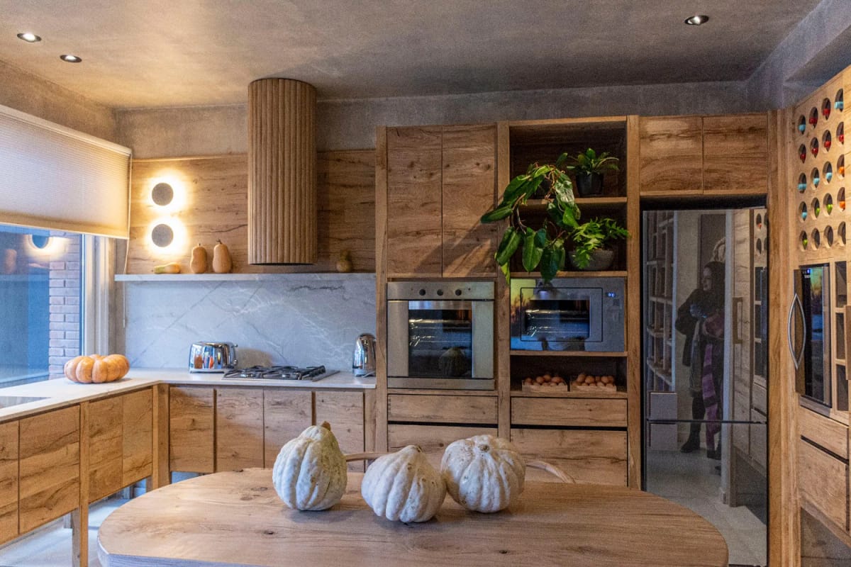 Japandi Kitchen Ideas: 10 Design Decisions That Keep It Warm, Not Clinical 14 A Japandi kitchen featuring natural wood cabinetry, a round wooden table with three white gourds, built-in ovens, open shelving with plants, and minimalistic decor that highlights warm textures instead of bright colors.