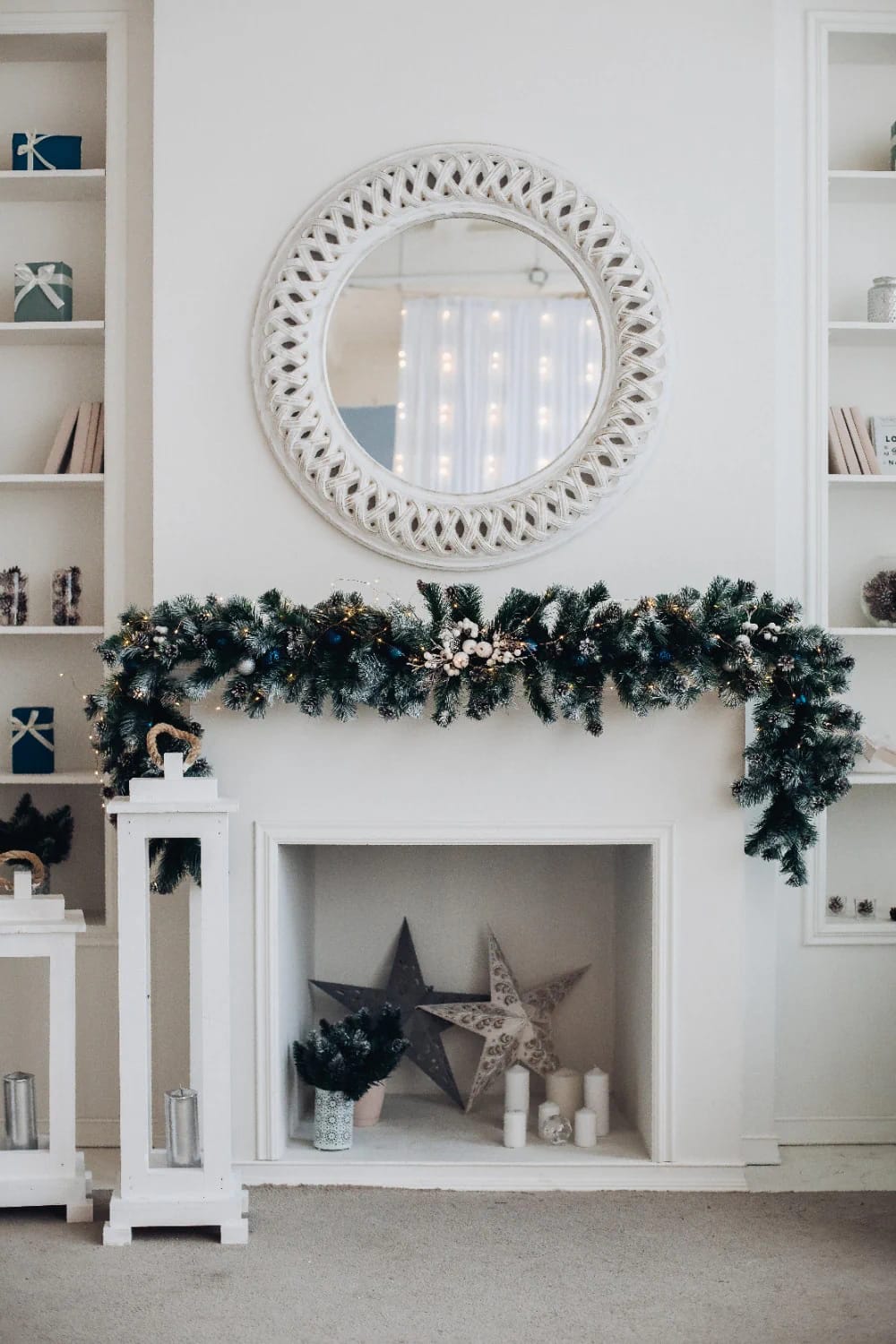 Blue and Silver Christmas Decor Ideas for the Living Room That Don't Read as Cold 10 A living room fireplace decorated with lush green garland featuring blue and silver accents, white candles, star ornaments, and a round white woven mirror above the mantel, illustrating blue and silver Christmas decor ideas for a cozy holiday look.