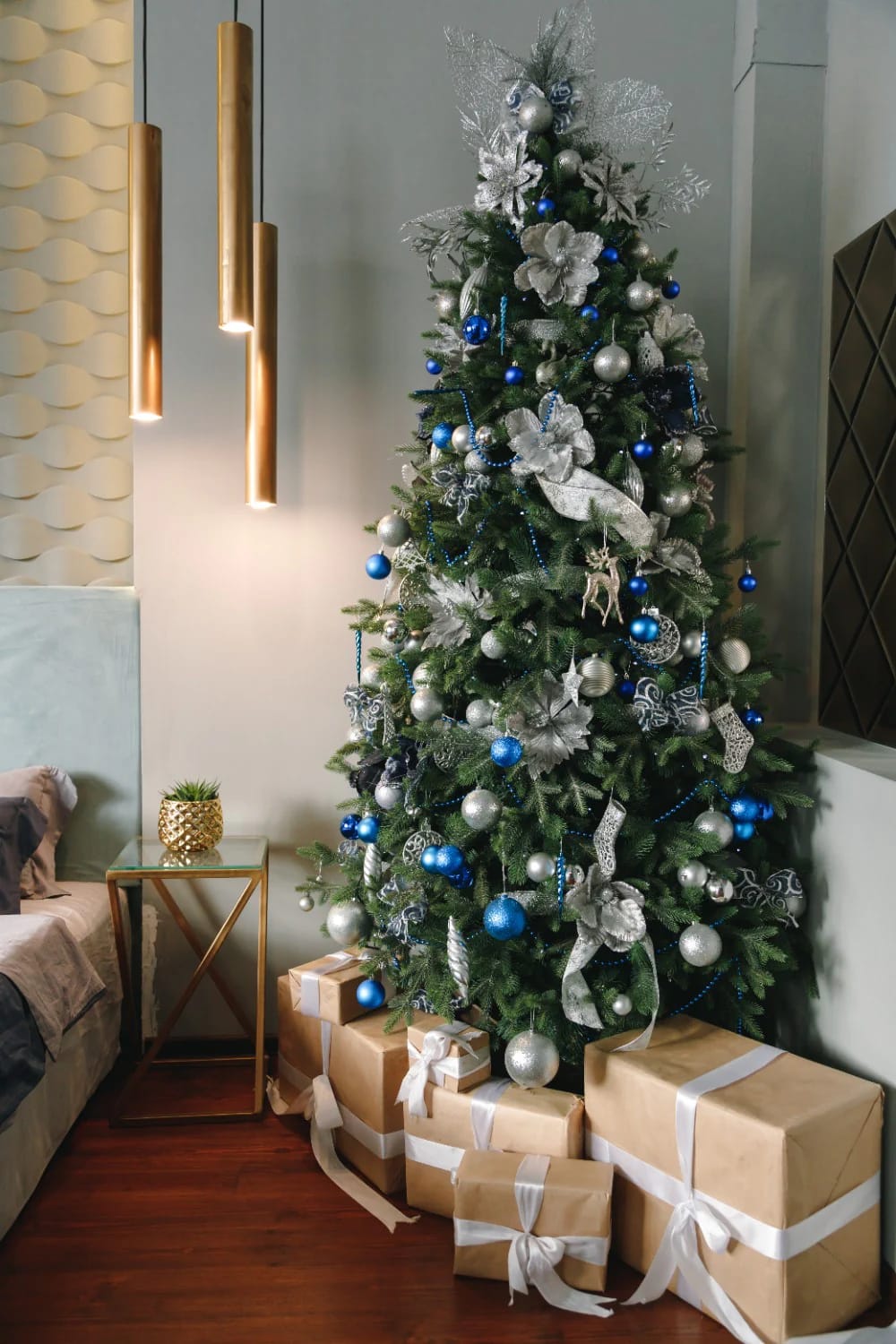 Blue and Silver Christmas Decor Ideas for the Living Room That Don't Read as Cold 3 A living room Christmas tree decorated with navy blue and silver ornaments, silver ribbons, and metallic floral accents, surrounded by gold-wrapped presents with white ribbons, illustrating blue and silver Christmas decor ideas that feel inviting rather than cold.