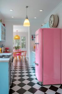 A vintage kitchen design featuring a pastel pink retro refrigerator, light blue cabinets, black and white checkered floor, and a dining area with orange chairs and a yellow pendant light, illustrating period-appropriate vintage kitchen decor.