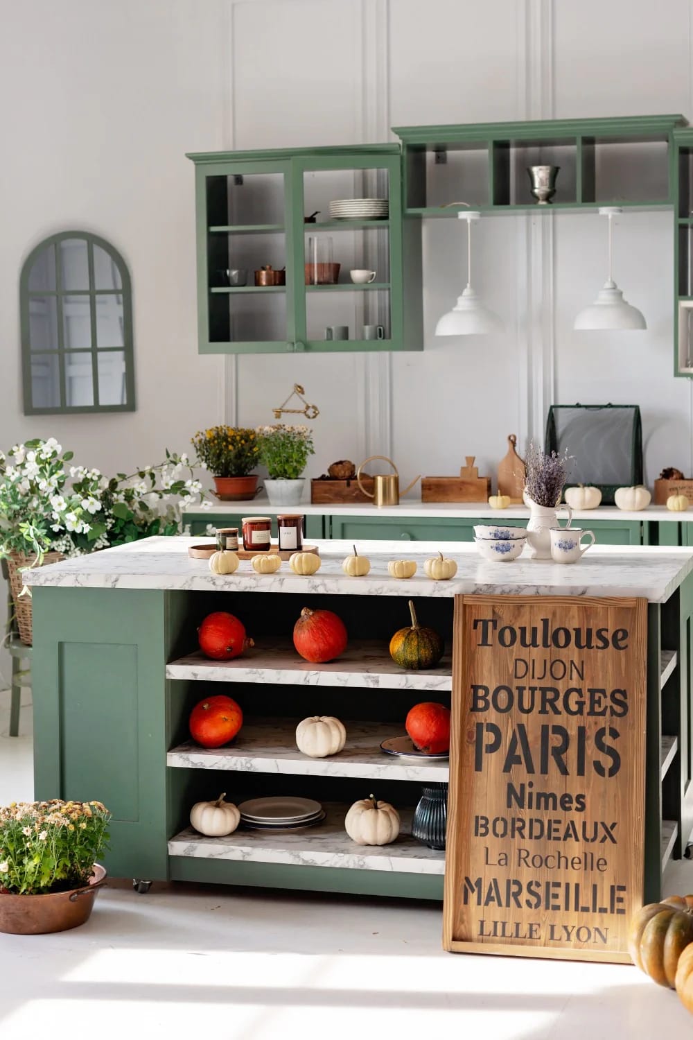 Cozy Kitchen Ideas That Actually Make You Want to Linger 7 A cozy kitchen featuring sage green cabinets, a marble countertop island with pumpkins and candles, open shelving with dishes, potted plants, and a decorative wooden sign, illustrating cozy kitchen ideas with sage green accents.