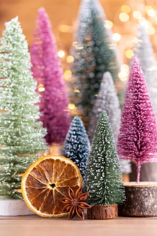 Whimsical Christmas Living Room Decor, Minus the Restraint 4 Miniature bottle brush Christmas trees in various pastel colors with snowy accents are arranged together on a wooden surface, accompanied by dried orange slices and a star anise, illustrating whimsical Christmas living room decor.