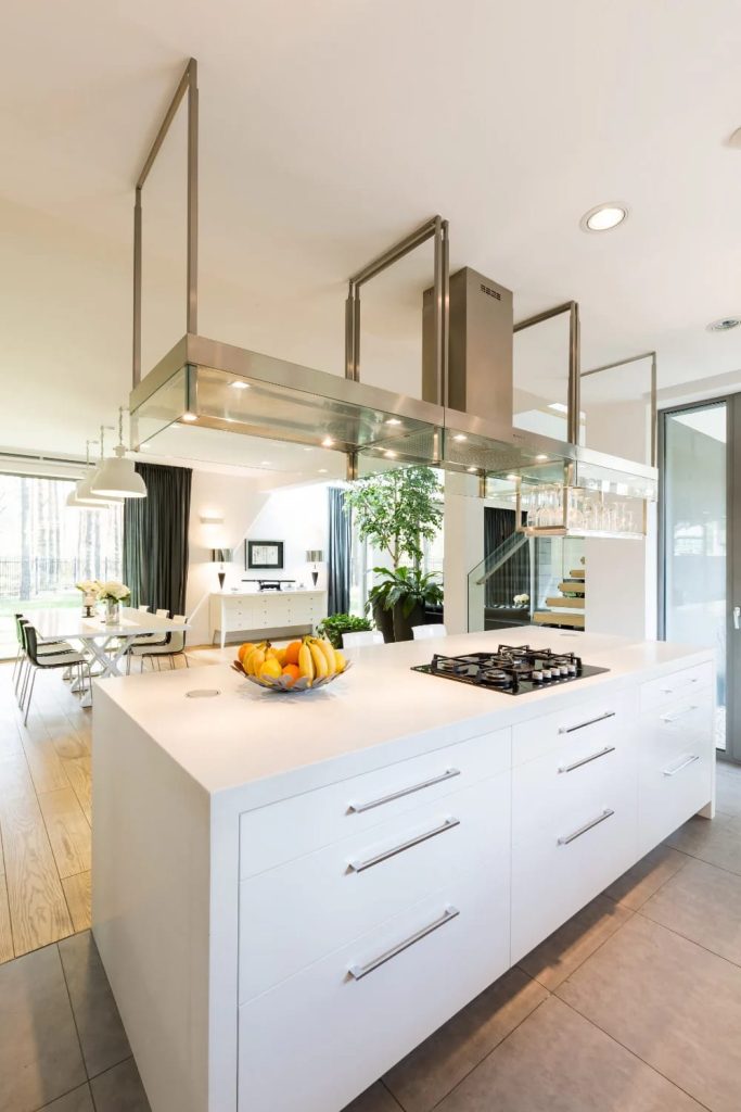Modern open concept floor plan featuring a bright kitchen island with a cooktop and fruit bowl, seamlessly connecting to a dining area and living space with large windows and indoor plants, illustrating the open concept vs closed floor plan article topic.