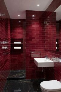 Modern bathroom featuring glossy burgundy subway tiles on the walls, black marble floor with white veining, a white floating sink, and a wall-mounted toilet, illustrating bold burgundy bathroom ideas with rich tile texture and sleek finishes.