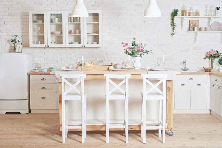Timeless Kitchen Ideas That Still Look Right in a Decade 1 A bright timeless kitchen featuring white cabinets, a wooden island with three white chairs, pendant lighting, glass-front cupboards, and subtle decorative touches like flowers and glassware, illustrating enduring kitchen design.