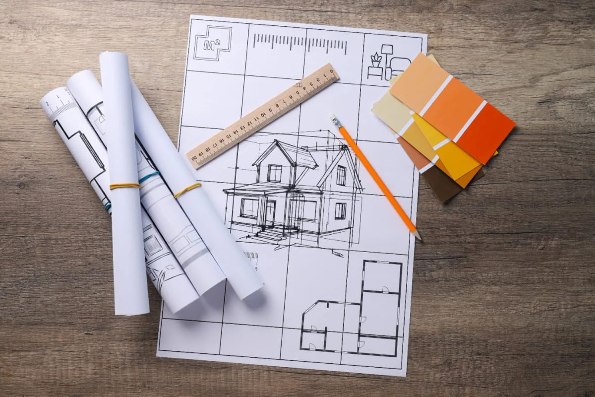 Architectural blueprints, a ruler, pencil, and color swatches on a wooden desk illustrating factors that make a floor plan expensive to build.