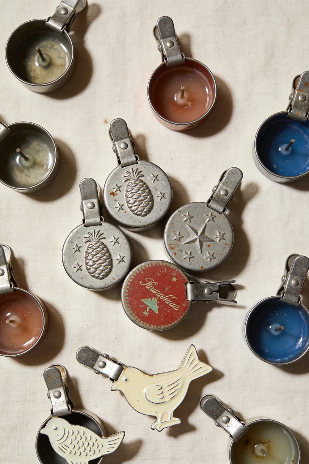 Assorted antique clip-on Christmas tree candles arranged on a light surface, including colored wax cups, metal lids with pineapple and star designs, and bird-shaped ornaments.
