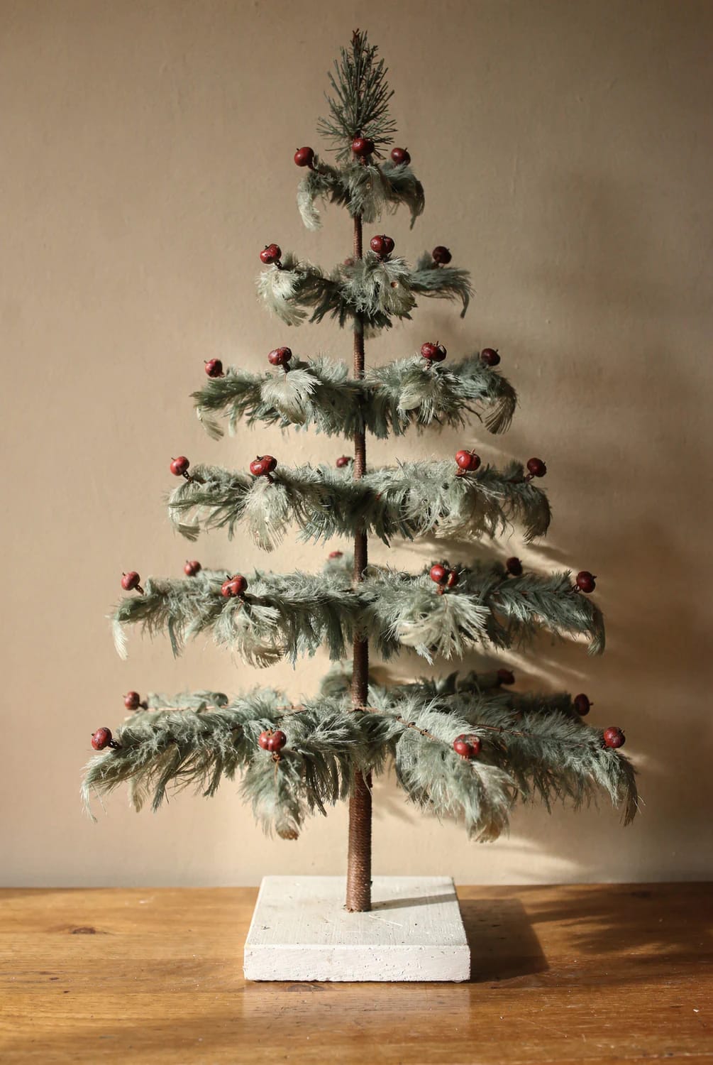 Antique feather Christmas tree with several tiered branches, red berry ornaments, and a white rectangular base displayed on a wooden table
