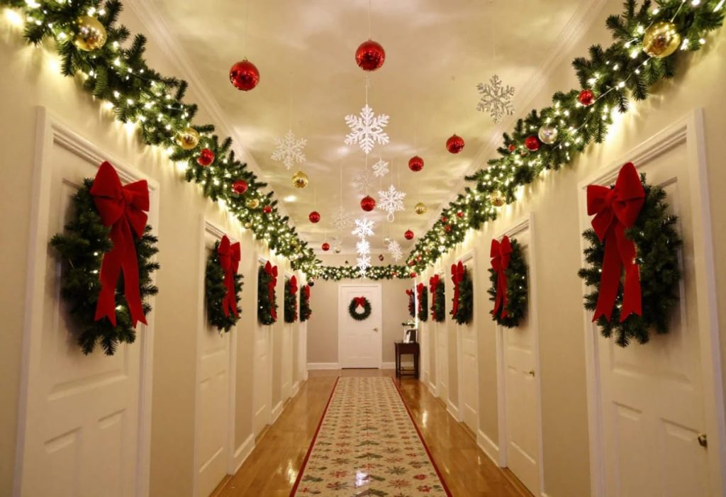 A narrow hallway decorated for Christmas with ceiling garlands, hanging ornaments, snowflake decorations, and wreaths with red bows on each door, showcasing festive hallway ceiling decor ideas for low ceilings.