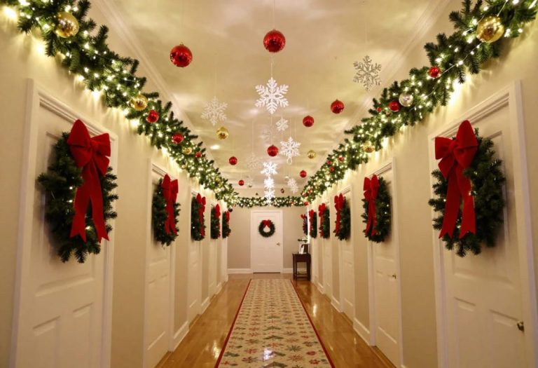 A narrow hallway decorated for Christmas with ceiling garlands, hanging ornaments, snowflake decorations, and wreaths with red bows on each door, showcasing festive hallway ceiling decor ideas for low ceilings.