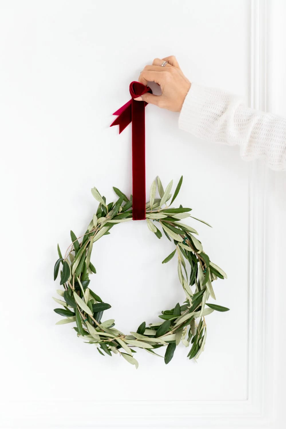 A hand hangs a simple green wreath with a red velvet ribbon on a white wall, demonstrating a Christmas hallway ceiling decor idea suitable for narrow halls and low ceilings.