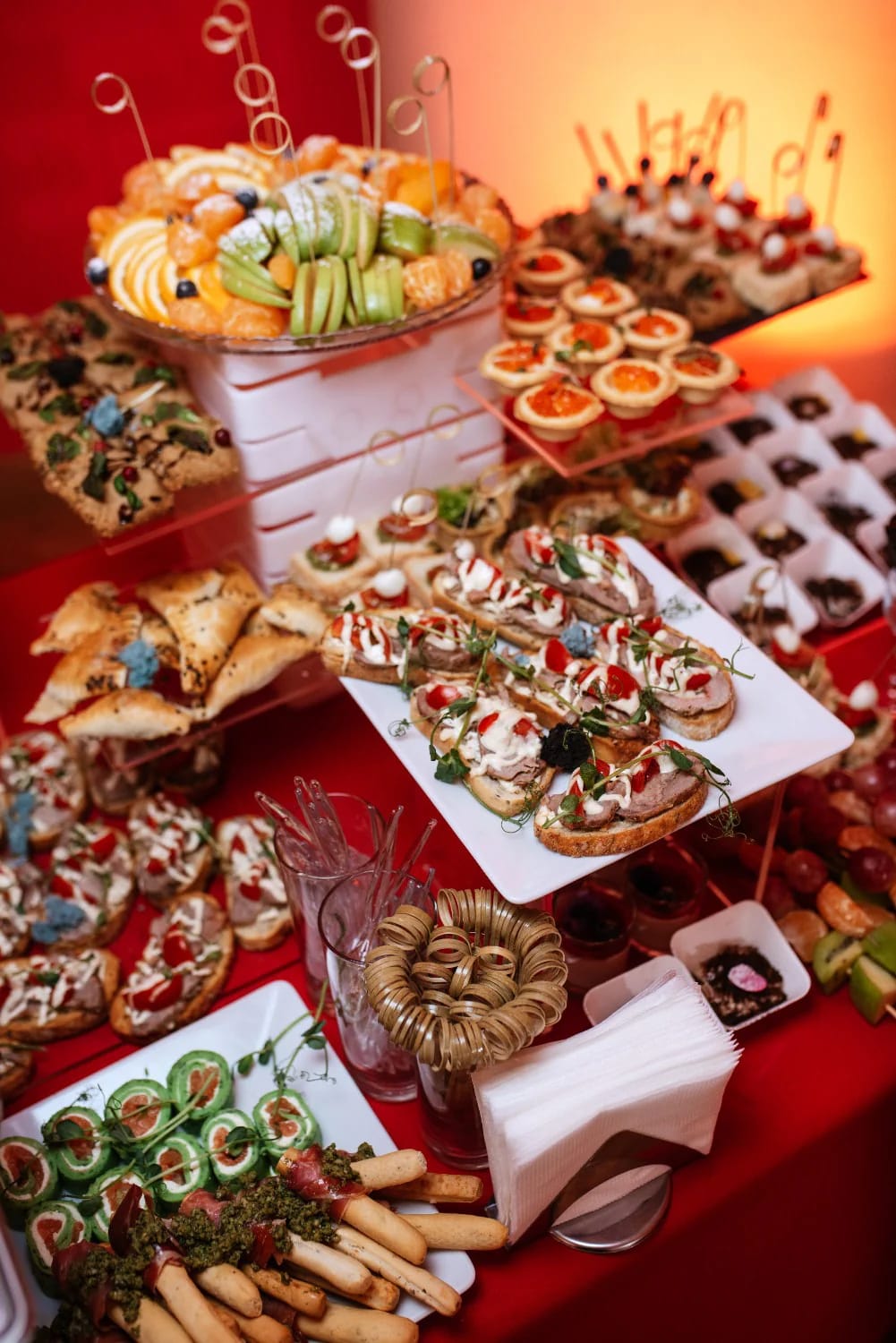 A Christmas party buffet table decorated with an assortment of savory appetizers, including fruit skewers, crostini, pastry triangles, breadsticks, and various bite-sized snacks, all arranged on tiered platters for a festive holiday presentation.