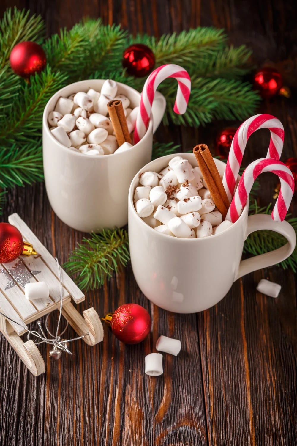 Two white mugs filled with hot cocoa topped with marshmallows, candy canes, and cinnamon sticks, surrounded by evergreen branches, red ornaments, and a small wooden sled, illustrating Christmas party dessert table decorating ideas.