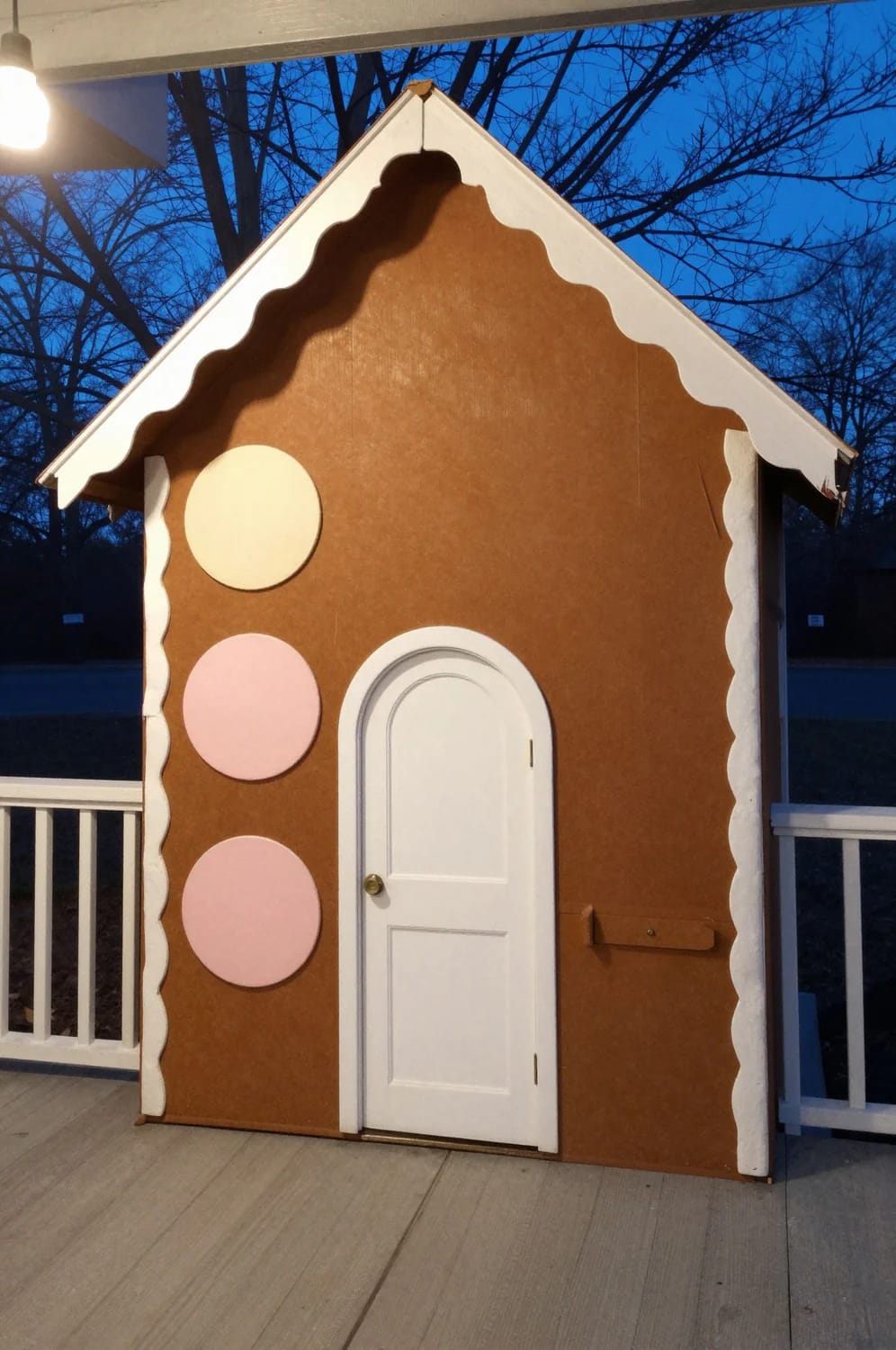 DIY Faux Gingerbread House Porch Facade A porch-scale DIY faux gingerbread house facade stands outdoors, featuring a brown front with white scalloped trim, pink and cream circular decorations, and a white arched door.