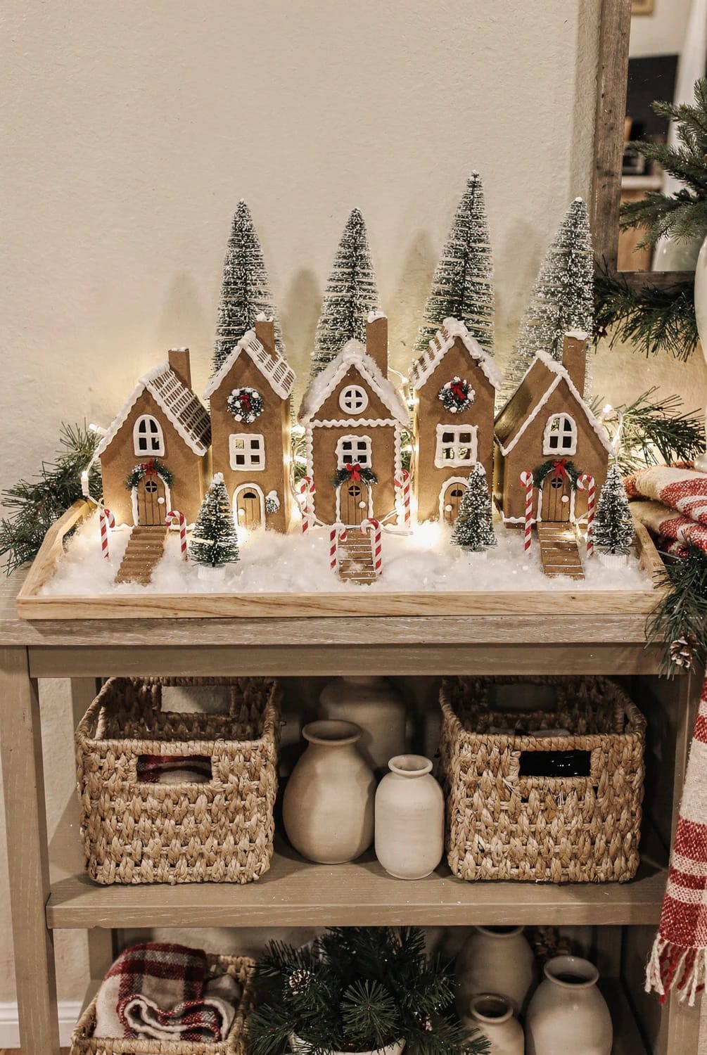 DIY Faux Gingerbread House Village Display A faux gingerbread village with five decorated houses, bottlebrush trees, candy canes, and warm lights arranged on a wooden console table for a seasonal display