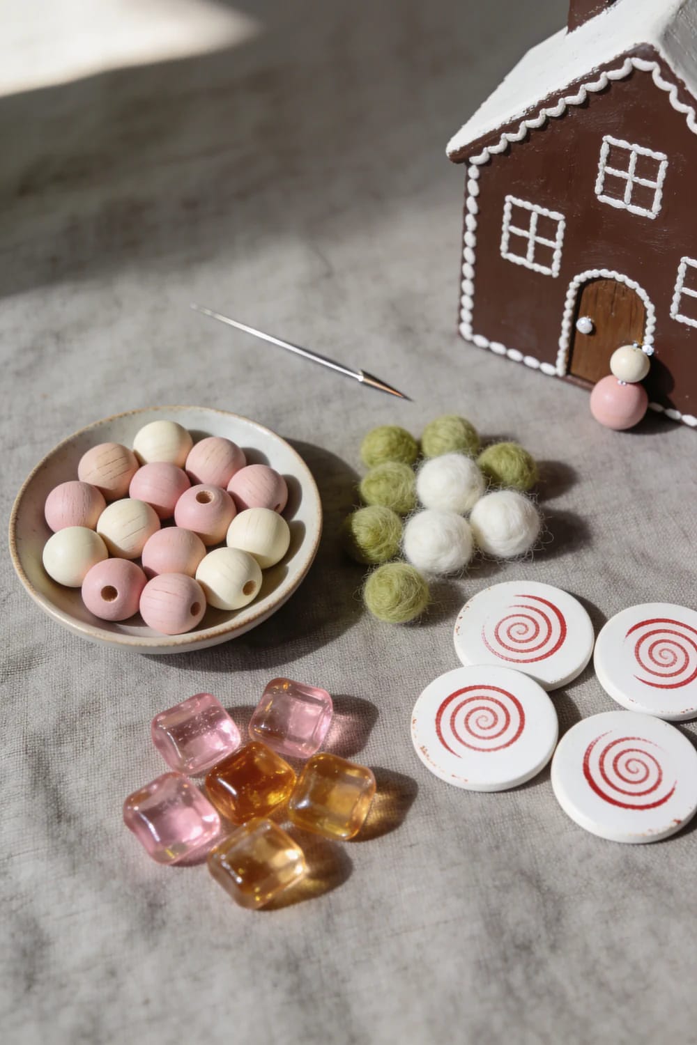 DIY Faux Gingerbread House Village Display A brown faux gingerbread house with white icing details sits beside wooden beads, felt balls, spiral discs, and translucent decorative pieces for a fake candy gingerbread village display