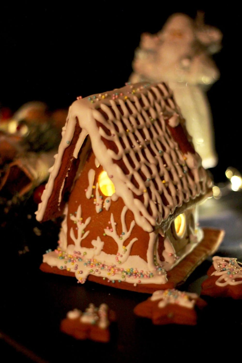 DIY Faux Gingerbread House Village Display Decorated gingerbread house with white icing, colorful sprinkles, and a warm light glowing through its front opening, with smaller gingerbread pieces around it in a dark display