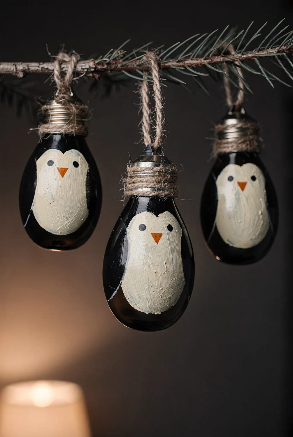 Three black painted light bulb penguin Christmas decorations hang from a pine branch by twine wrapped around their screw caps.