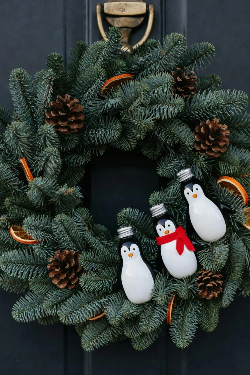 Evergreen Christmas wreath decorated with pinecones, dried orange slices, and three penguin ornaments made from light bulbs, including one wearing a red scarf