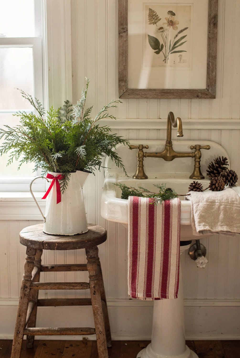Farmhouse Christmas Bathroom Cedar Pitcher Decor A white enamel pitcher filled with cedar clippings and tied with a red ribbon sits on a rustic wooden stool beside a pedestal bathroom sink, with pinecones and striped towels adding farmhouse Christmas decor