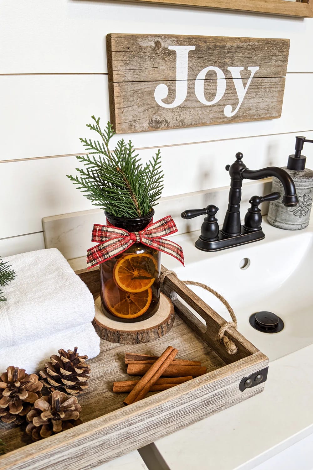 Farmhouse Christmas Bathroom Dried Orange Decor Farmhouse Christmas bathroom with a jar of dried orange slices and cedar greenery tied with a plaid bow, displayed on a wood tray beside a sink with pinecones and cinnamon sticks.