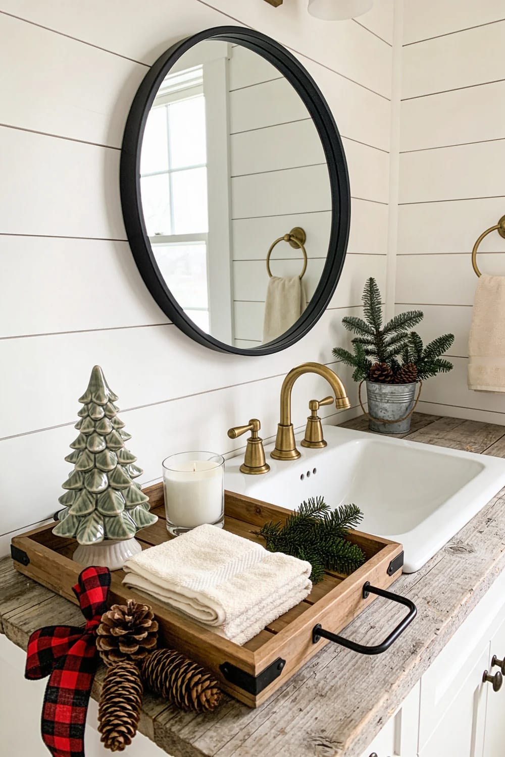 Farmhouse Christmas Bathroom Vanity Tray Farmhouse Christmas bathroom vanity with a wooden tray holding folded white towels, a ceramic tree, candle, pine branches, pinecones, and a red plaid ribbon beside a white sink and oval black-framed mirror