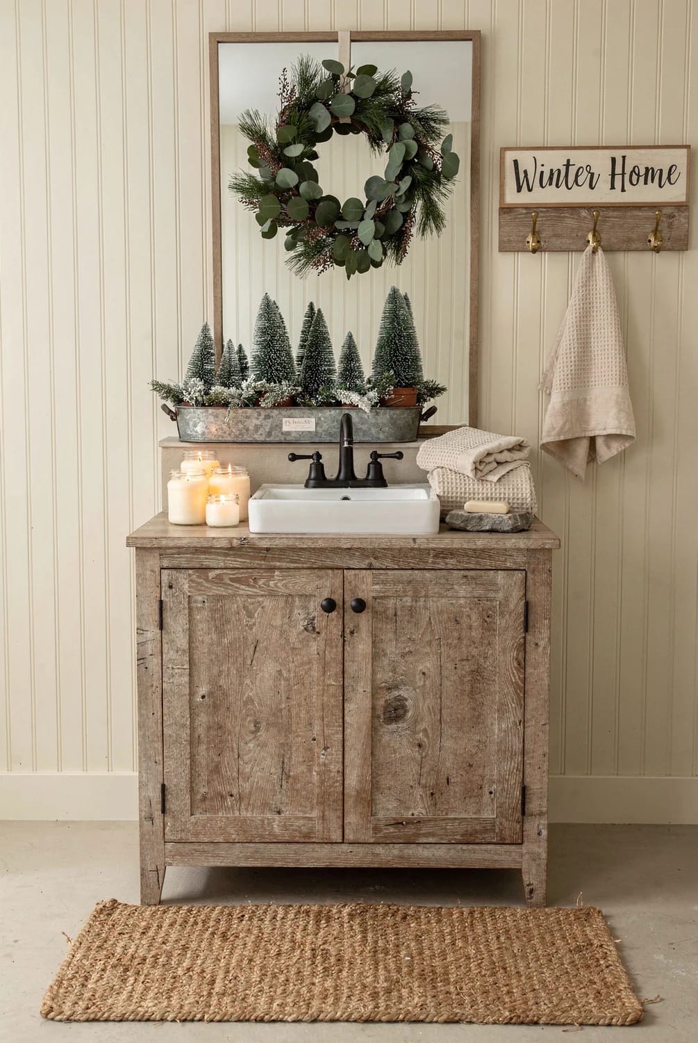 Farmhouse Christmas Bathroom With Greenery Farmhouse Christmas bathroom featuring a weathered wood vanity, white sink, eucalyptus and evergreen wreath, miniature bottle-brush trees in a metal tray, cream towels, candles, and a Winter Home wall sign