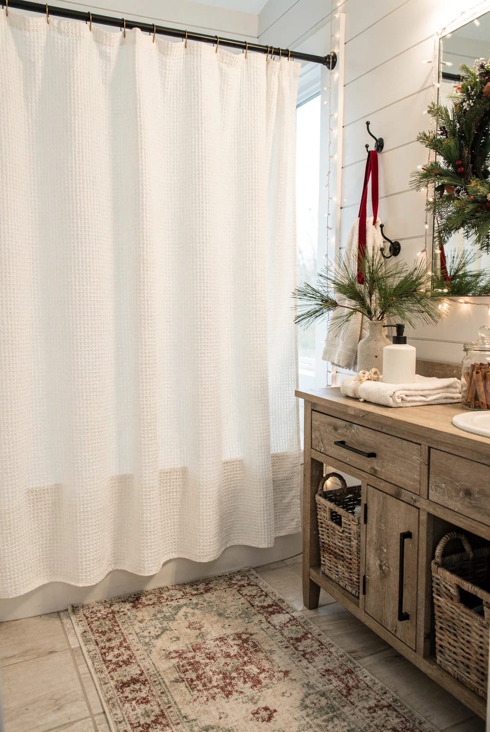 Farmhouse Christmas Waffle-Weave Shower Curtain Farmhouse Christmas bathroom featuring a white waffle-weave shower curtain, rustic wood vanity, woven baskets, red towels, evergreen branches, and string lights