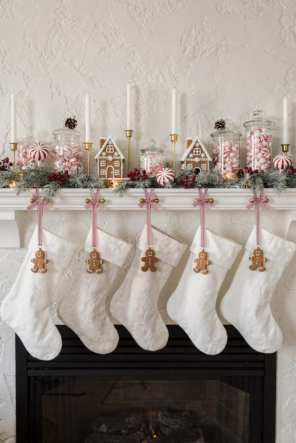 Christmas mantel decorated with gingerbread houses, red-and-white candy jars, striped ornaments, evergreen garland, candles, and five stockings with gingerbread man ornaments