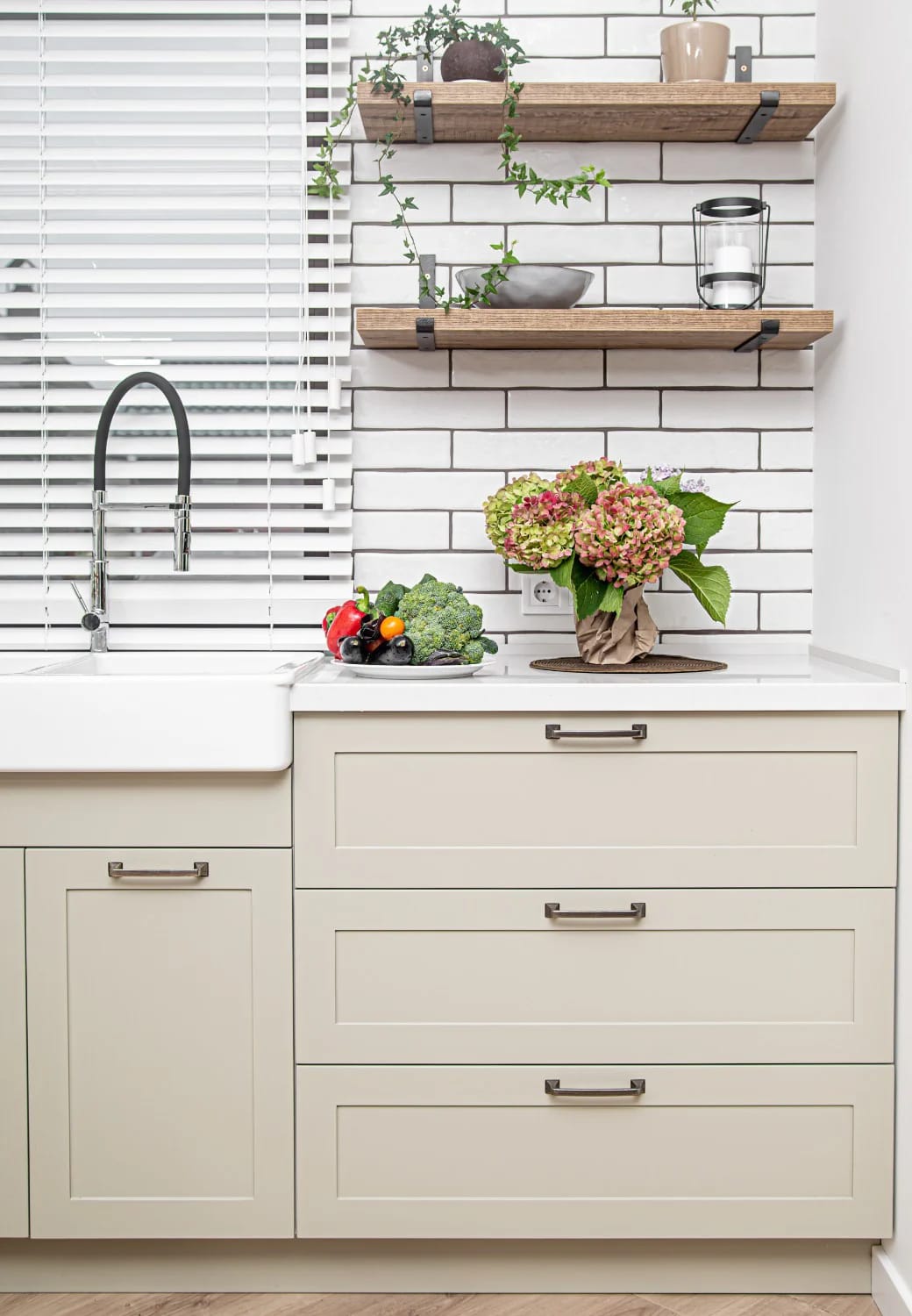 Light Colored Kitchen Cabinets: 7 Ways to Get Bright Without Going Cold 13 Modern kitchen with light greige and taupe cabinets, a white farmhouse sink, subway tile backsplash, wooden shelves with plants and decor, and a countertop featuring fresh vegetables and a floral arrangement, illustrating bright kitchen cabinet ideas.