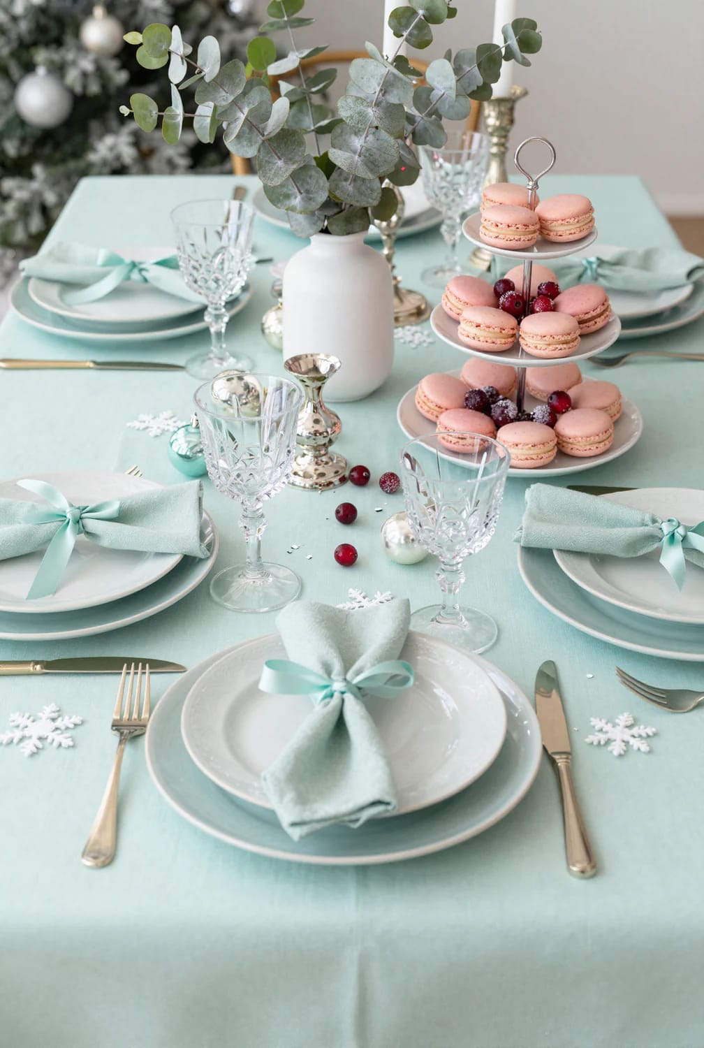 Pastel Pink and Mint Christmas Decorations That Still Look Like Christmas 23 A festive dining table decorated with pastel pink and mint Christmas decorations, featuring a mint tablecloth, pink macarons on a tiered tray, white plates with mint napkins tied with blue ribbons, crystal glasses, silver cutlery, snowflake ornaments, and a white vase with eucalyptus branches.
