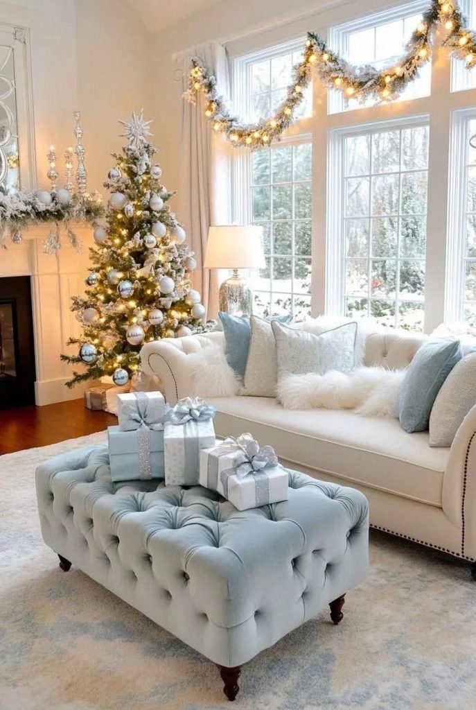 Living room decorated with icy blue and white winter wonderland Christmas decor, featuring a Christmas tree with white and blue ornaments, a garland with warm 2700K lights, a tufted blue ottoman with wrapped gifts, and a cream sofa with blue and white pillows, with no ceiling lights visible.
