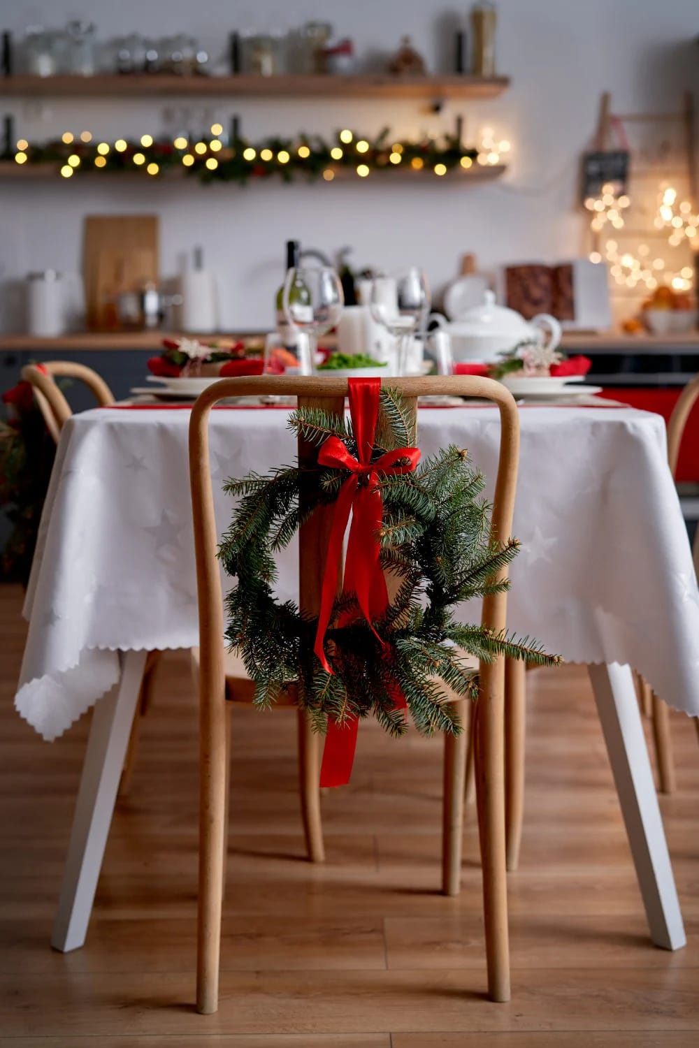 A dining chair decorated with a green wreath and a red ribbon bow, showcasing red ribbon Christmas decor beyond the tree, in a festive dining room setting with a white tablecloth and holiday lights in the background.