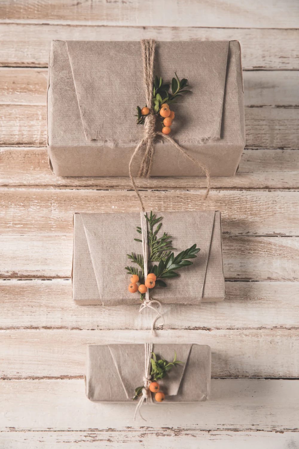 Scandinavian Christmas Decor With Pale Wood and Straw Ornaments That Reads Nordic, Not Rustic 22 Three gifts wrapped in kraft paper and tied with string, each decorated with small sprigs of greenery and orange berries, displayed on a pale wood surface to reflect Scandinavian Christmas decor style.