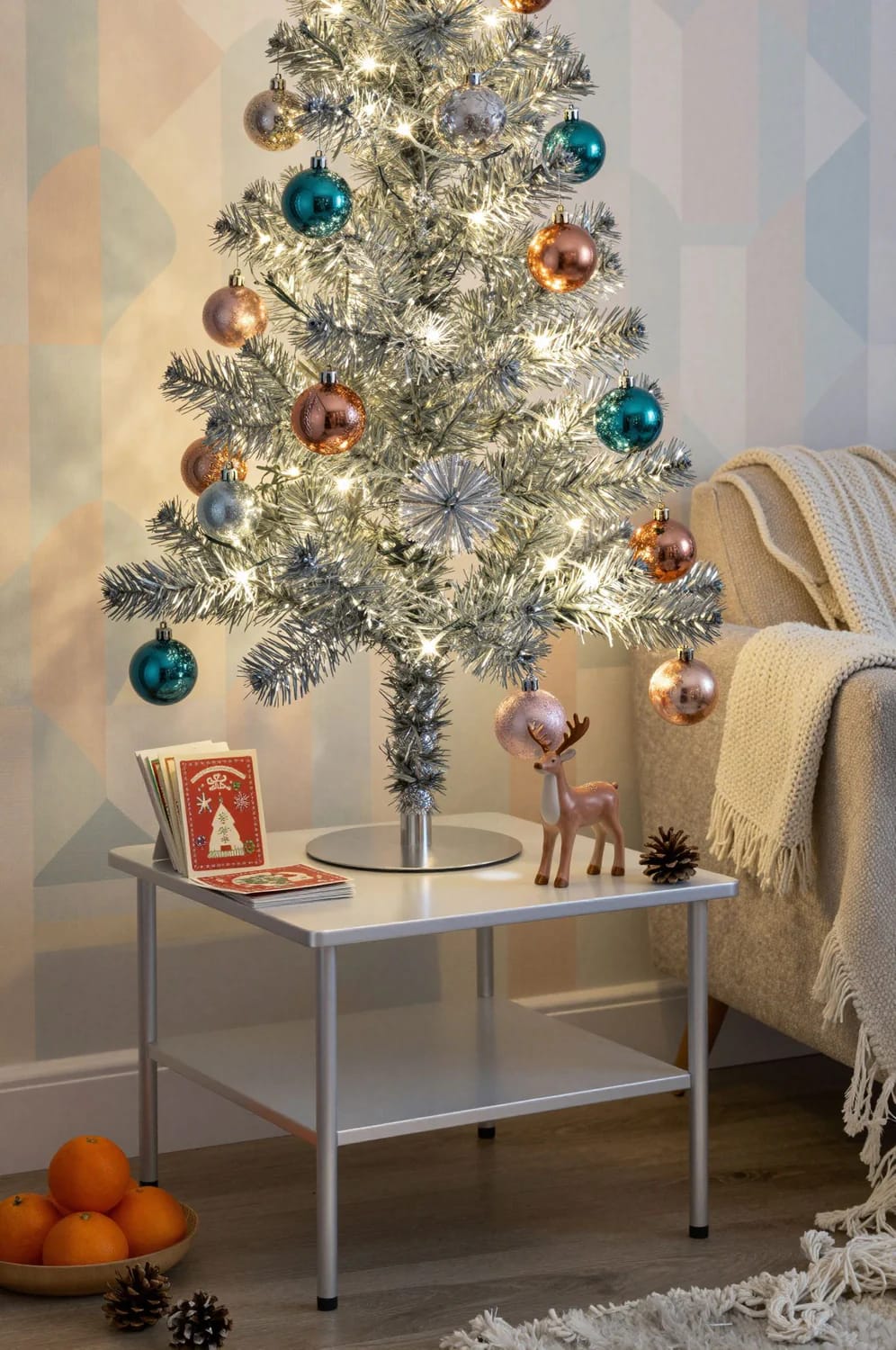 Small Apartment Christmas Tree Ideas for a Slim or Tabletop Tree That Doesn't Look Like a Compromise 18 A slim aluminum tabletop Christmas tree decorated with colorful ornaments and white lights sits on a small table beside a cozy sofa, with holiday cards, a reindeer figurine, pine cones, and oranges arranged nearby, illustrating small apartment Christmas tree ideas.
