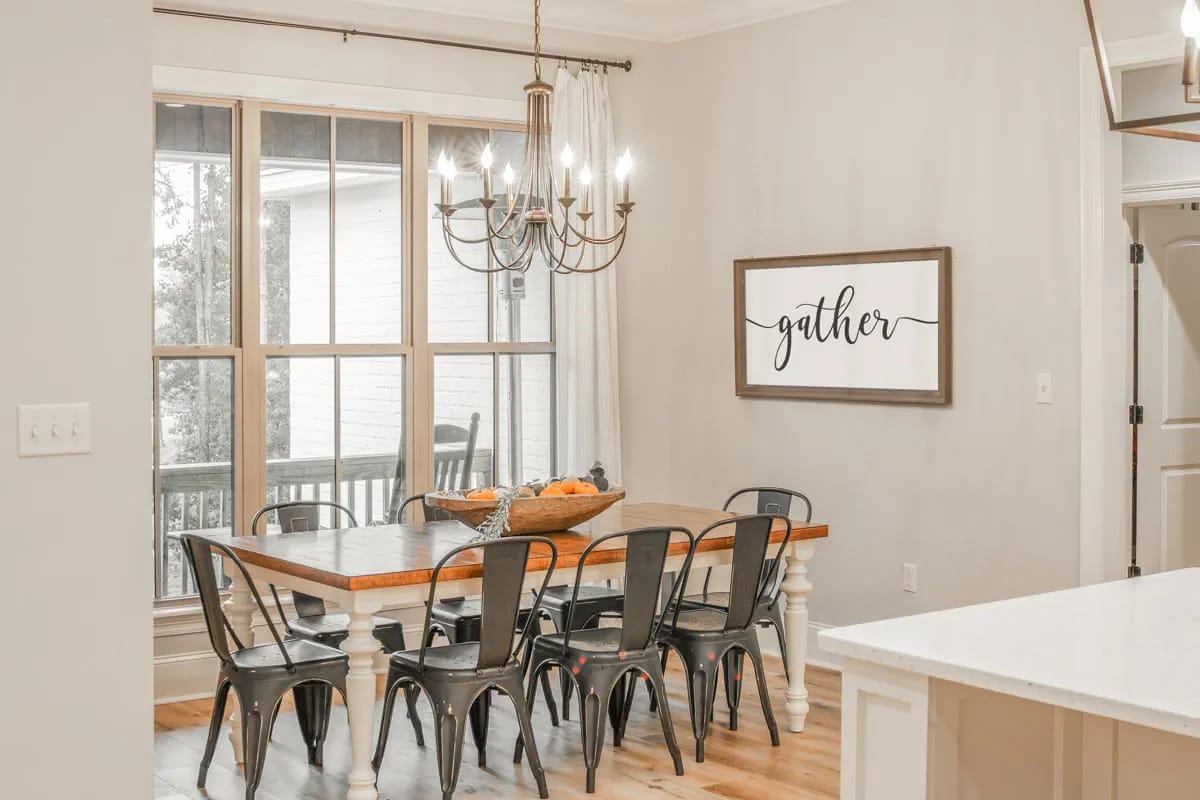 Bright dining area with wood table, metal chairs, chandelier, and tall windows overlooking a covered outdoor space