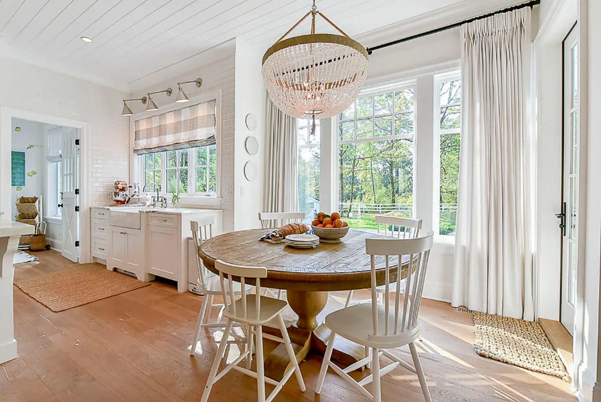 Bright dining nook with round wooden table, white chairs, and large windows beside a farmhouse kitchen
