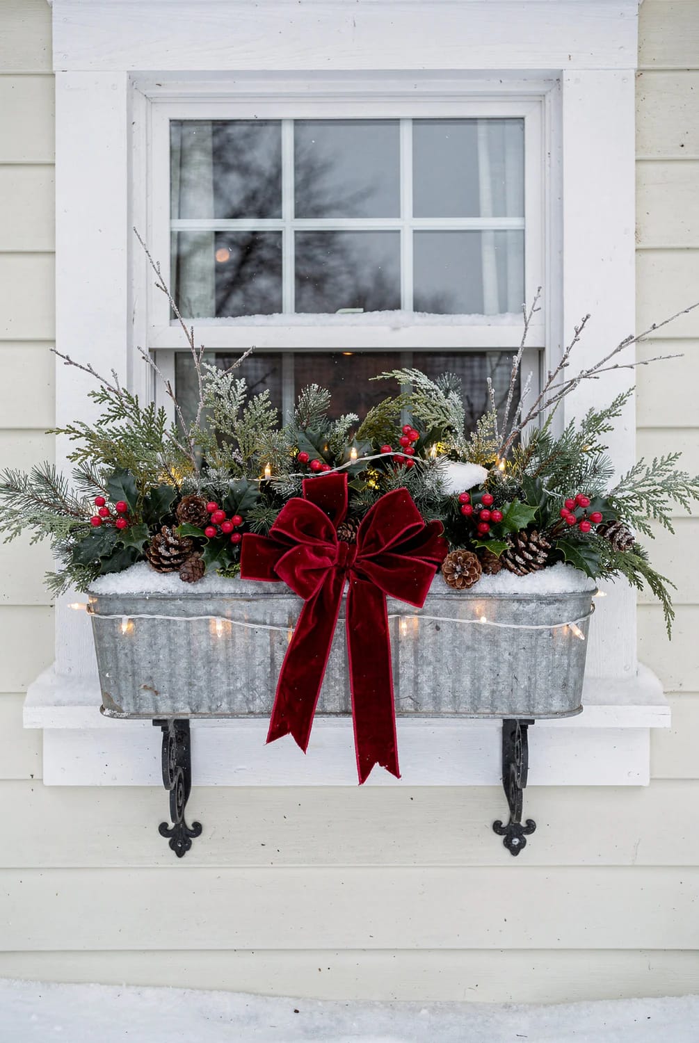 Christmas window box decorating idea featuring a galvanized trough mounted beneath a window with evergreen branches, red berries, pinecones, snow, warm string lights, and a large burgundy bow