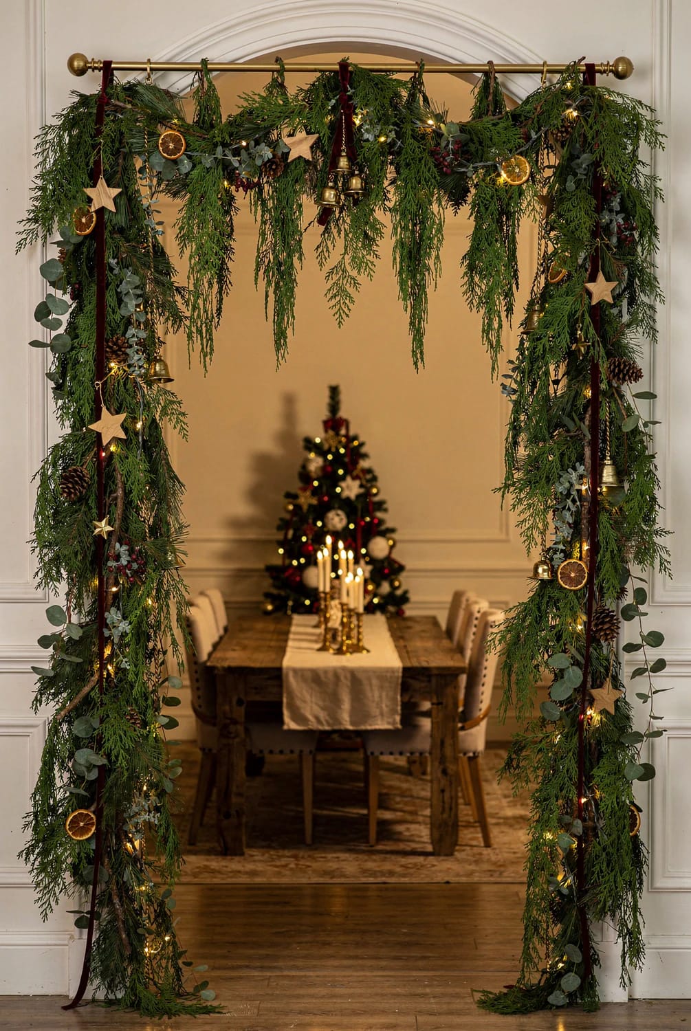 A decorated interior doorway is framed with evergreen Christmas garland, while multiple vertical strands hang from a brass rod like a curtain. Warm string lights, dried orange slices, wooden stars, bells, eucalyptus, and pinecones decorate the garland.