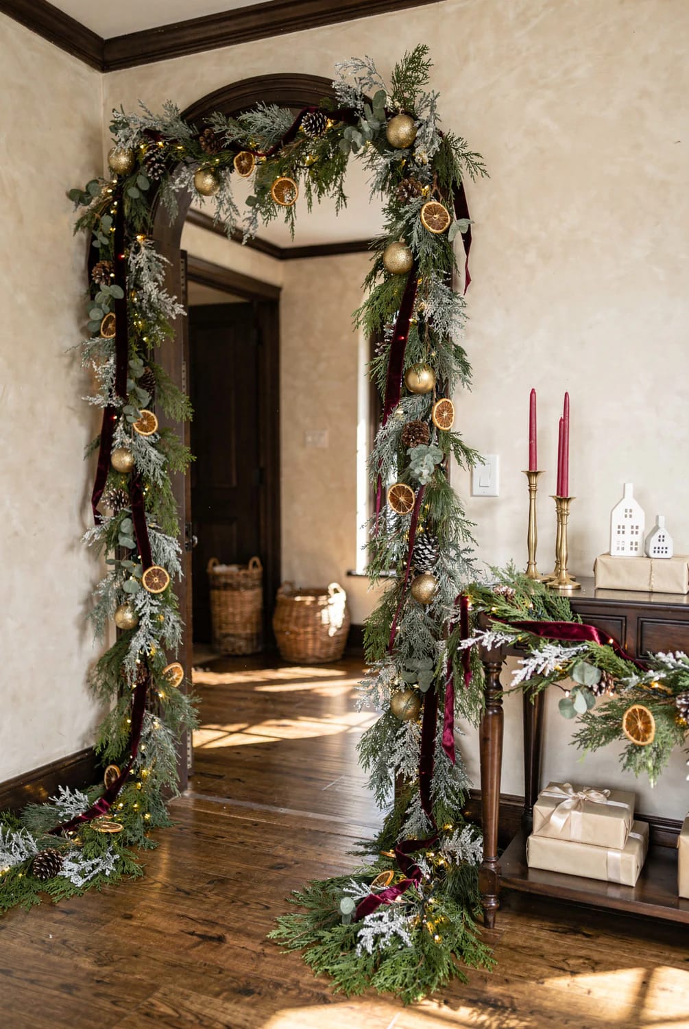 Christmas garland decorates an interior archway with evergreen branches, frosted foliage, dried orange slices, pinecones, gold ornaments, lights, and burgundy ribbon extending to the floor and pooling on the wood floor