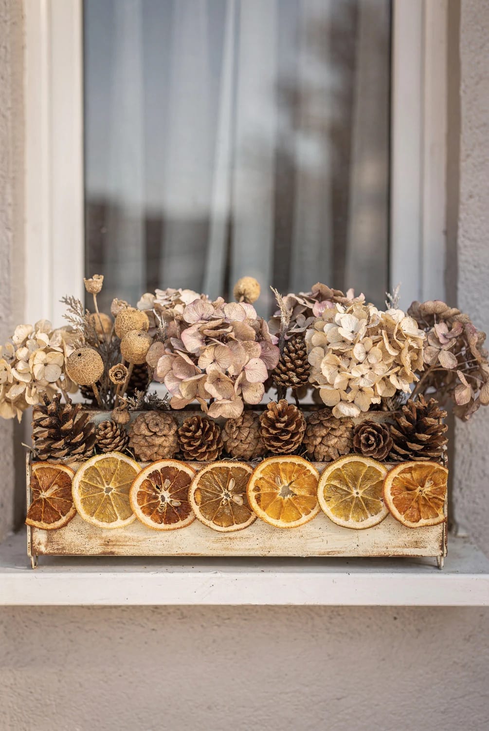 Christmas window box filled with dried hydrangea heads, pinecones, seed pods, and sliced dried oranges arranged on an exterior windowsill