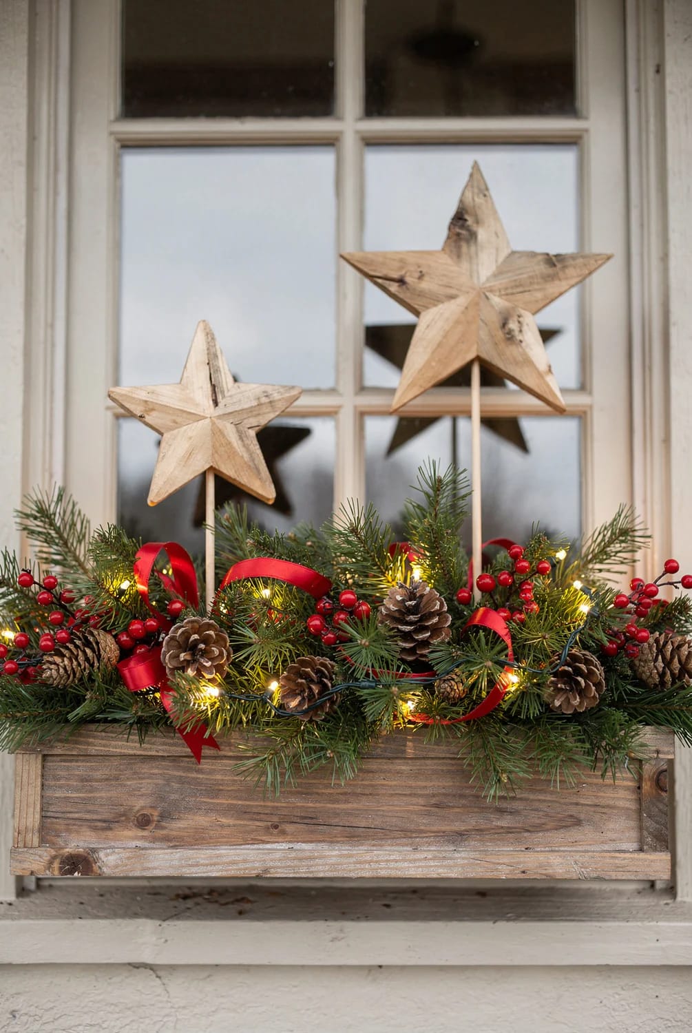12 Christmas Window Box and Exterior Sill Decorating Ideas That Survive January 3 Christmas window box filled with evergreen branches, red berries, pinecones, red ribbon, and warm lights, featuring two wooden stars on stakes at different heights