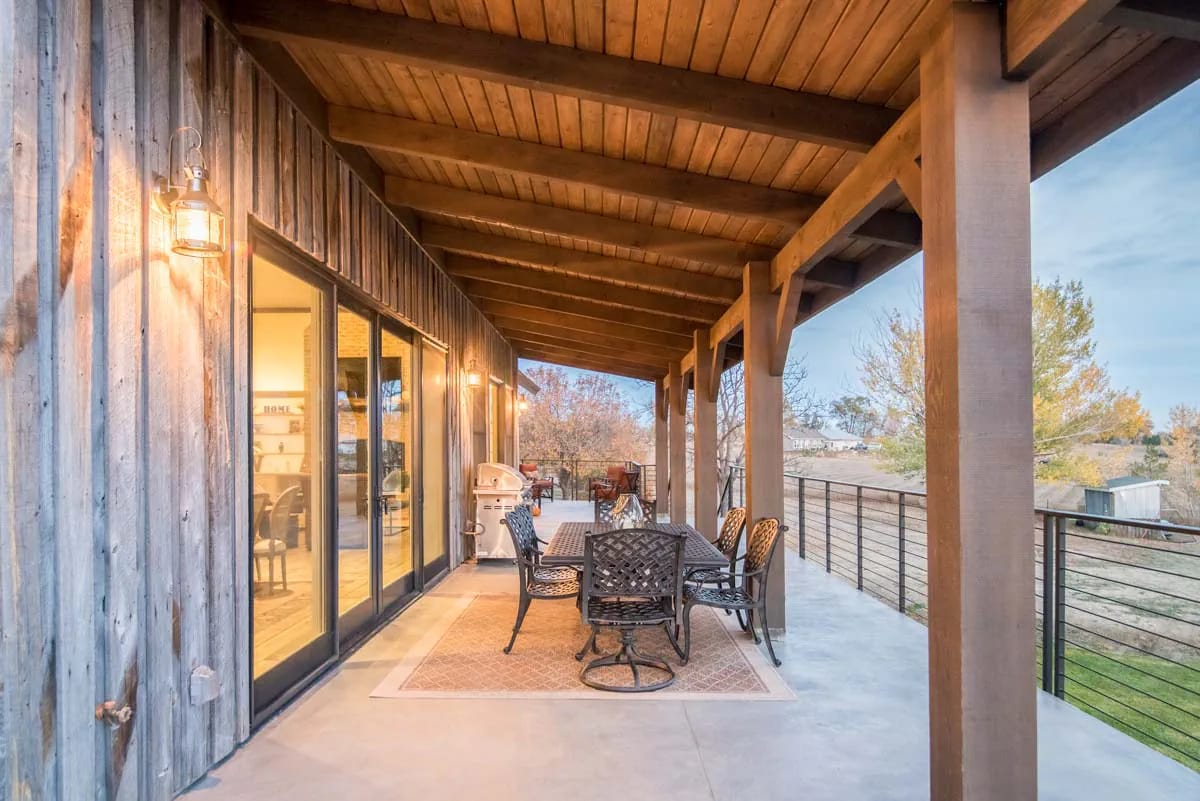 Covered porch with timber beams, rustic siding, outdoor dining set, and metal railing overlooking landscape.