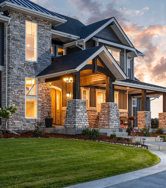 Craftsman-style home with stone and timber porch, large windows, and multiple gables at dusk