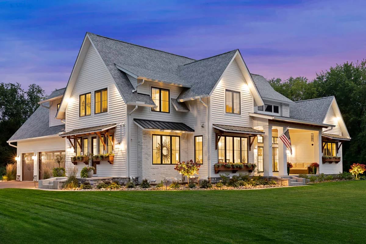 Dusk exterior of a large white-sided home with multiple gables, warm lighting, covered entry, and attached garage