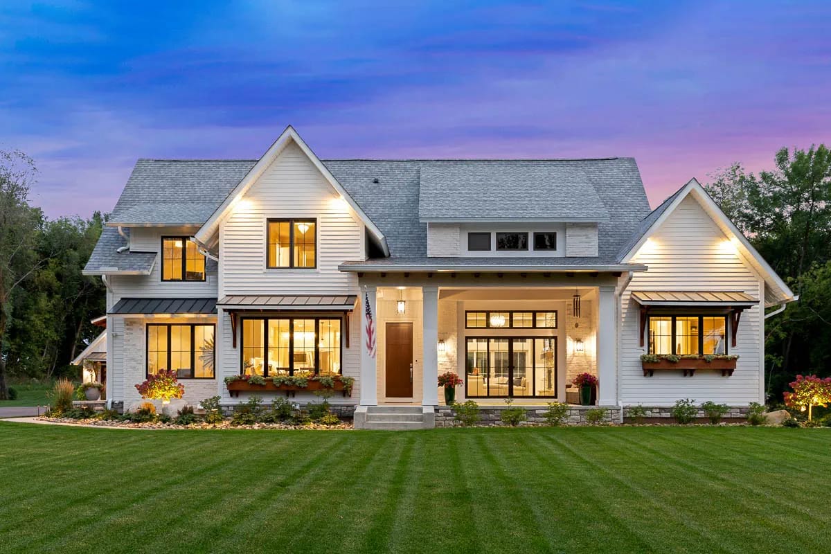 Dusk exterior view of a white gabled home with stone accents, illuminated windows, and a covered central entry