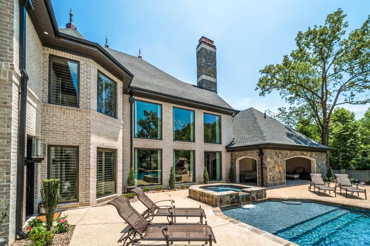Elegant rear exterior with brick and stone, large windows, pool, and covered patio